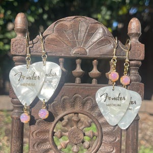 May include: Earrings featuring pearlescent white guitar picks with the "Fender" logo and "Medium" text. Each earring has a gold-colored hook and a small, iridescent pink bead. The earrings are displayed on a wooden stand.