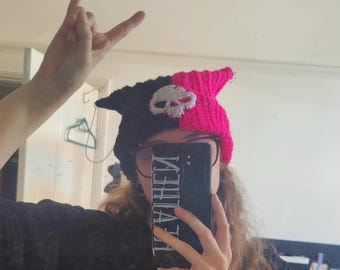 Puede incluir: Una persona lleva una gorra de punto negra y rosa con un diseño de calavera y la palabra "HEAVEN" escrita en una funda de teléfono negra.
