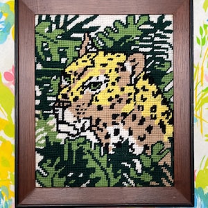 May include: A framed needlepoint artwork featuring a leopard amidst green foliage. The leopard has yellow and brown spots, with a tan face and white accents. The frame is a dark brown wood, and the background is a floral pattern.