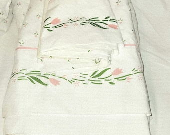 May include: A folded white bed sheet set featuring a repeating pattern of small pink flower buds and a prominent horizontal border design of green leaves and pink tulips, accented by a thin pink horizontal line across the fabric surface.
