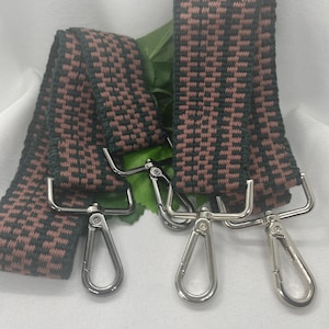 May include: Two woven straps with a brown and black checkered pattern. Each strap has silver-colored metal swivel clasps attached. The straps are displayed on a white surface with green leaves.