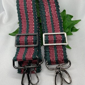 May include: Two woven bag straps with a striped pattern in dark green and burgundy. Each strap has metal hardware, including adjustable buckles and swivel clasps. The straps are displayed against a white background with green leaves.
