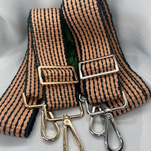 May include: Two striped purse straps with gold and silver hardware. The straps feature a peach and black woven pattern. Each strap has a metal buckle and a swivel clasp for attachment.