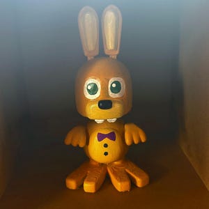 May include: A handcrafted, orange-yellow bunny figurine with large, upright ears and a small purple bow tie. The bunny has green eyes, a black mouth, and three black buttons. The figurine is in a cardboard box.