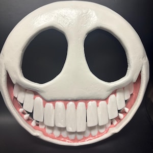 May include: A white, smiling mask with large, rectangular teeth and black eye holes. The mask has a wide grin with pink gums. The mask is a full face design.