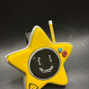 May include: A yellow star-shaped device with a black screen displaying a smiling face made of white dots. It has a small antenna and two colored buttons. A small triangle with a pattern is on the bottom left. The device is on a black surface.