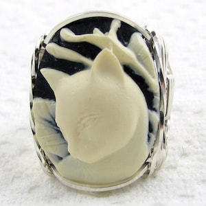 May include: A silver ring featuring a black and white cameo of a cat's head. The cameo is set in a silver bezel and the ring band is decorated with a braided design.