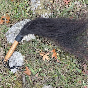 Puede incluir: Un matamoscas de crin de caballo negro con un mango de madera marrón claro. El mango está sujeto a la crin de caballo con un cordón negro envuelto. Un cordón negro está sujeto al extremo del mango. El matamoscas está sobre la hierba.