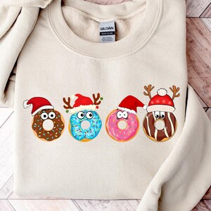 Könnte beinhalten: Ein beiger Pullover mit einem weihnachtlichen Design mit vier Donuts mit Gesichtern und Weihnachtsmützen. Die Donuts sind mit Streuseln und Zuckerguss verziert.
