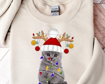 May include: A beige sweatshirt with a gray cat wearing a red Santa hat and reindeer antlers with Christmas lights. The cat is surrounded by colorful Christmas lights.