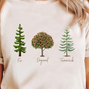 Può includere: Una maglietta beige con una grafica di tre alberi: un abete, un corniolo e un larice. Gli alberi sono illustrati in stile acquerello. Il testo "Fir", "Dogwood" e "Tamarack" è stampato sotto ogni albero.
