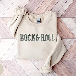 Puede incluir: Una sudadera de color arena con el texto "ROCK & ROLL" impreso en una fuente verde oscuro y desgastada.