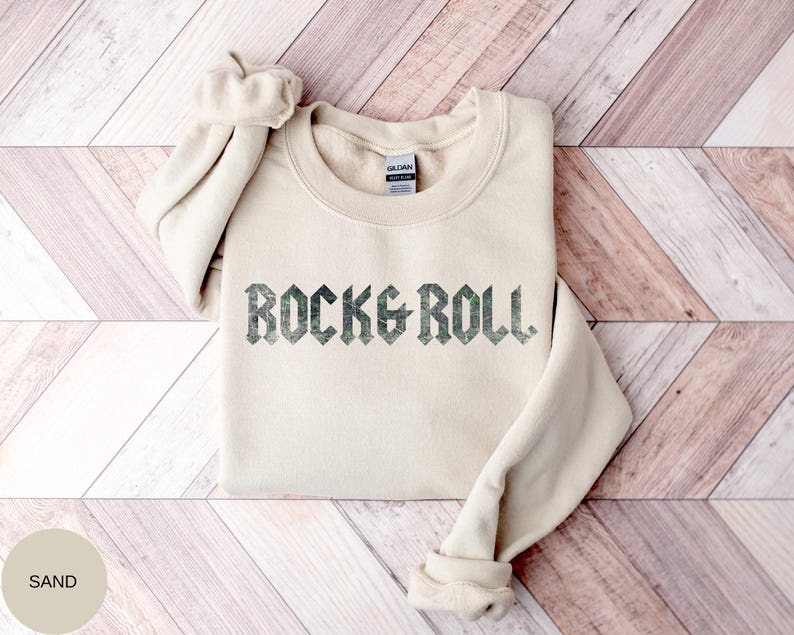 Puede incluir: Una sudadera de color arena con el texto "ROCK & ROLL" impreso en una fuente verde oscuro y desgastada.