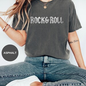 Puede incluir: Una mujer lleva una camiseta gris oscuro con las palabras "Rock & Roll" impresas en purpurina plateada. Lleva pantalones vaqueros azules y zapatillas blancas.