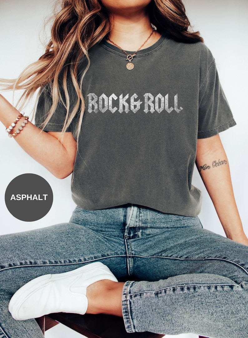 Puede incluir: Una mujer lleva una camiseta gris oscuro con las palabras "Rock & Roll" impresas en purpurina plateada. Lleva pantalones vaqueros azules y zapatillas blancas.