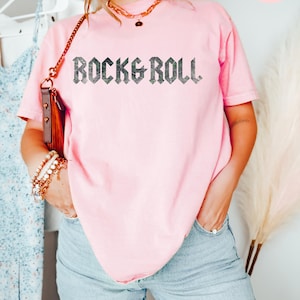 Puede incluir: Una camiseta rosa claro con el texto "Rock & Roll" impreso en negro en la parte delantera. El texto est&aacute; en una fuente g&oacute;tica en negrita.