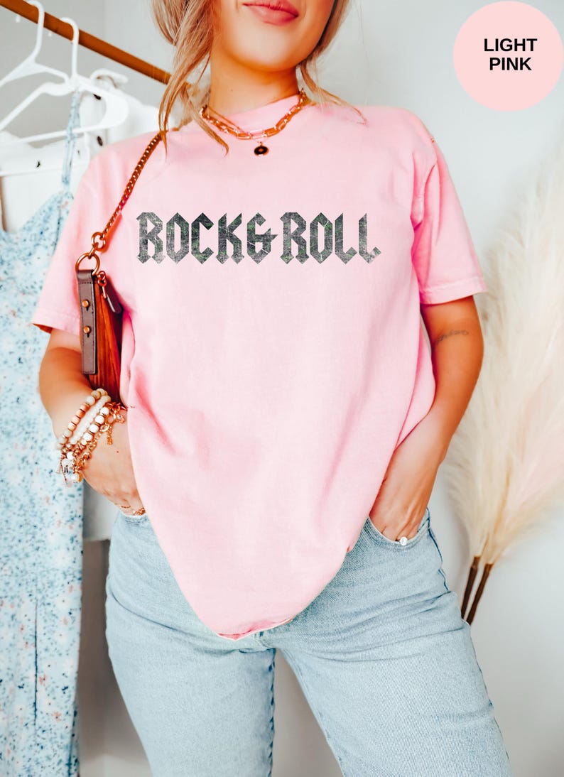 Puede incluir: Una camiseta rosa claro con el texto "Rock & Roll" impreso en negro en la parte delantera. El texto est&aacute; en una fuente g&oacute;tica en negrita.