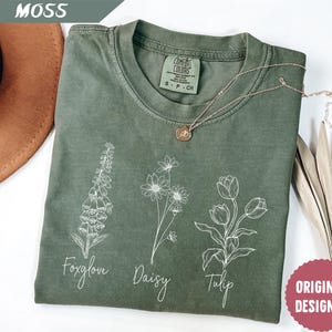 Może przedstawiać: Zielona koszulka z białym obrysem naparstnicy, stokrotki i tulipana. Koszulka ma napisane słowa "Foxglove", "Daisy" i "Tulip" pod każdym kwiatem. Koszulka ma również napis "COMFORT COLORS" i "ORIGINAL DESIGNER".