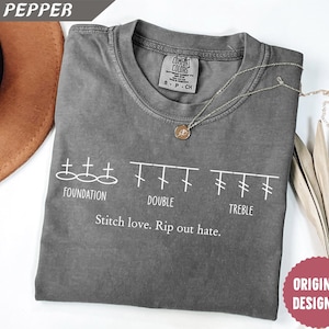 May include: A grey Comfort Colors t-shirt with white text reading "FOUNDATION, DOUBLE, TREBLE" and "Stitch love. Rip out hate." A gold necklace with a pendant rests on the shirt. The shirt is folded.