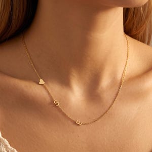 Può includere: Una collana color oro caratterizzata da una catenina sottile con un piccolo ciondolo a forma di cuore e le lettere S ed E distanziate lungo il filo. La collana poggia sul collo, mettendo in risalto un design essenziale con elementi in metallo lucido.