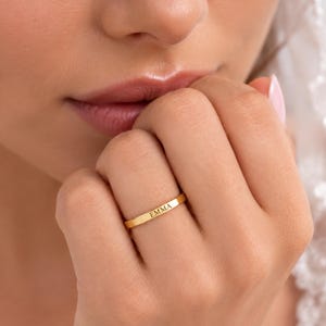 Könnte beinhalten: Ein dünner goldfarbener Ring mit dem eingravierten Namen EMMA in einer deutlichen, dunklen Schrift auf der Außenseite. Der Ring hat eine glatte, polierte Oberfläche und ein flaches Profil und wird an einem Finger neben weißem Spitzenstoff getragen.