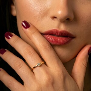 Peut inclure: Une bague en or avec une petite pierre précieuse ronde et transparente. La bague est portée au doigt d'une femme, avec du vernis à ongles rouge foncé. Les lèvres de la femme sont rouges et sa peau est claire.