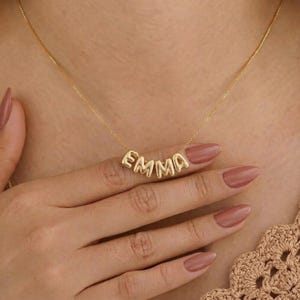 Könnte beinhalten: Eine goldfarbene Halskette mit dem Namen "EMMA" in einzelnen, abgerundeten Buchstaben. Die Halskette wird um den Hals getragen. Die Buchstaben sind durch eine zarte Kette verbunden. Die Halskette hat eine warme Goldfarbe.