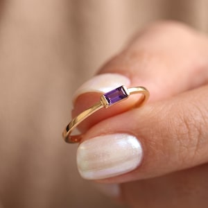 May include: A thin gold ring featuring a single rectangular baguette-cut purple amethyst gemstone set horizontally. The band is smooth and polished, showcasing a minimalist design that highlights the vibrant violet hue of the central stone.