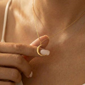Può includere: Una collana con pendente a forma di mezzaluna dorata. La collana presenta una delicata catena e un ciondolo a forma di luna curva e lucida. Il gioiello è esposto su uno sfondo color carne.