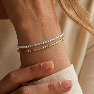 Könnte beinhalten: Zwei zarte Armbänder am Handgelenk. Ein Armband ist silberfarben und zeigt ein Muster aus kleinen runden Perlen und kurzen zylindrischen Stäbchen. Das zweite Armband ist goldfarben und weist ein identisches Muster aus Perlen und Stäbchen auf.
