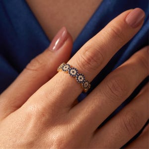 May include: A gold band ring featuring a continuous row of circular evil eye motifs. Each motif is crafted with a central white stone surrounded by a ring of small blue stones, creating a repeating pattern around the entire circumference of the band.