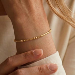 Puede incluir: Una delicada pulsera de tono dorado que presenta un patrón repetitivo de pequeñas cuentas redondas y finas barras cilíndricas. La pieza de joyería minimalista está diseñada con una estructura de cadena delgada que rodea la muñeca.