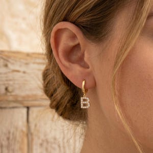 May include: A gold hoop earring featuring a dangling letter B charm. The letter B is encrusted with small, clear, sparkling stones that cover its entire front surface. The hoop is a simple, polished gold band that secures the letter charm in place.