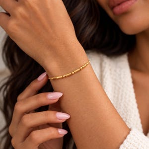 Puede incluir: Una delicada pulsera de tono dorado que presenta una secuencia de pequeñas cuentas esféricas lisas intercaladas con cuentas tubulares cilíndricas alargadas, creando un patrón minimalista que rodea la muñeca con un acabado pulido y brillante.