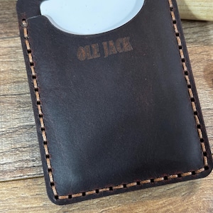 May include: A dark brown leather cardholder with the text "OLE JACK" embossed on the front. The cardholder has a stitched border with light brown thread and a curved top opening to reveal a white card.