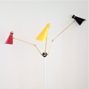 May include: A mid-century modern floor lamp with three adjustable arms, each holding a cone-shaped shade. The shades are red, yellow, and black. The lamp has a white pole and a black circular base, offering a stylish lighting solution.