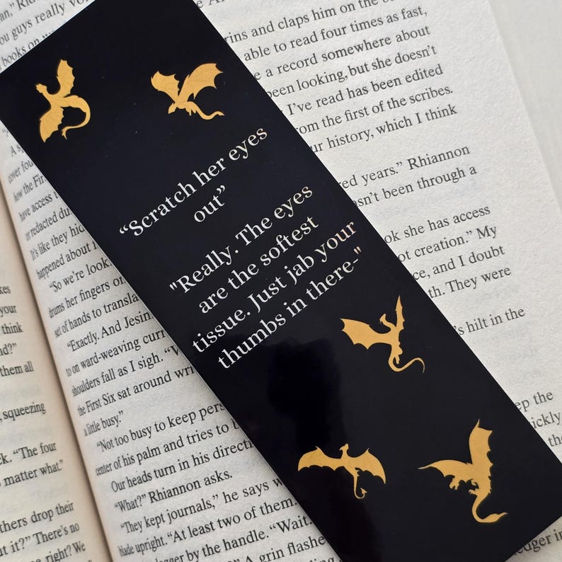 May include: Black bookmark featuring gold dragon silhouettes and the text "Scratch her eyes out" and "Really. The eyes are the softest tissue. Just jab your thumbs in there - "