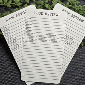 May include: Three white rectangular book review cards with rounded corners, featuring black printed text and lines. Each card includes fields for title, author, series, genre, format, start date, finished date, and ratings for overall, world building, plot, spice, character, and ending, followed by lined space for thoughts.