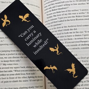 May include: Black bookmark featuring gold dragon silhouettes and the quote "Can you carry a luminary while insulted?" printed in white.