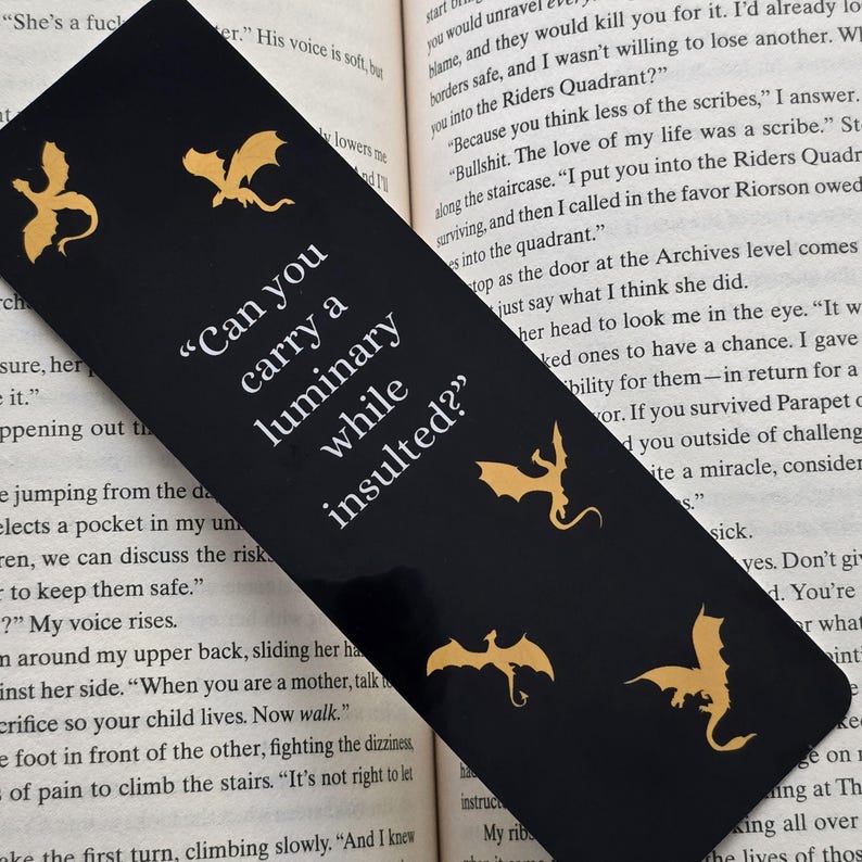 May include: Black bookmark featuring gold dragon silhouettes and the quote "Can you carry a luminary while insulted?" printed in white.