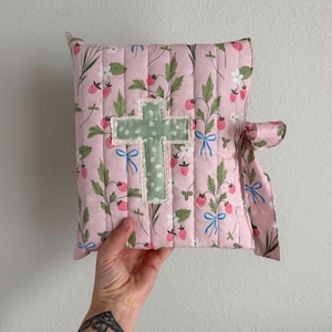May include: A pink quilted Bible cover with a strawberry and floral pattern. A green cross with white lace trim is centered on the cover. Blue bows accent the design, and a pink ribbon secures the cover.