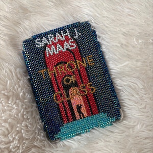 Puede incluir: Una funda de teléfono adornada con pedrería, que muestra la portada del libro "Throne of Glass" de Sarah J. Maas. La funda está adornada con cristales azules, rojos y dorados. El título y el nombre del autor son visibles.