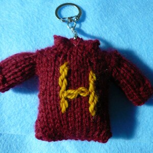 May include: A maroon knitted sweater keychain with a yellow embroidered letter "H" on the front. The keychain has a silver key ring attached.