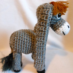 May include: A gray crocheted donkey with a brown mane and a white face. The donkey has black hooves and a long, black tail.