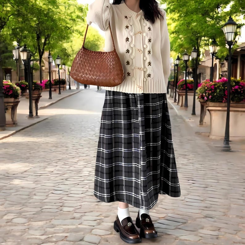 May include: A woman wearing a white cardigan with floral embroidery and a brown woven handbag. She is also wearing a black and white plaid skirt and brown loafers.
