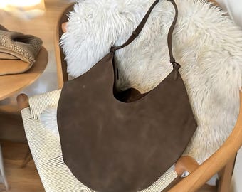 Peut inclure: Un sac à main en daim marron avec une seule bandoulière. Le sac a une forme arrondie en croissant et est posé sur une surface en fausse fourrure blanche.