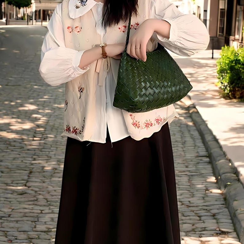 May include: A woman wearing a white blouse with floral embroidery and a dark green woven handbag.