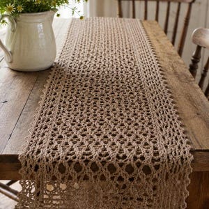 May include: A tan crochet table runner with a delicate openwork design. The runner is placed on a wooden table, with a white pitcher of flowers in the background. The runner's dimensions are approximately 60 inches long.