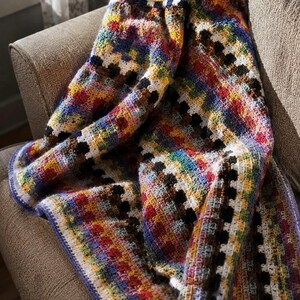 Peut inclure: Une couverture crochetée à la main, aux couleurs vives, drapée sur un fauteuil neutre. La couverture présente un motif patchwork avec des carrés de différentes couleurs, dont le rouge, le bleu, le jaune et le noir. Les bords sont finis avec une bordure bleue.