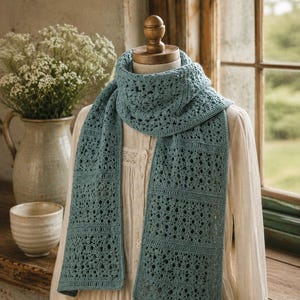 May include: A teal-colored, open-weave crochet scarf is draped on a mannequin. The scarf has a rectangular shape with a repeating pattern of small holes. The mannequin is wearing a cream-colored blouse. A vase of white flowers and a small ceramic cup are in the background.